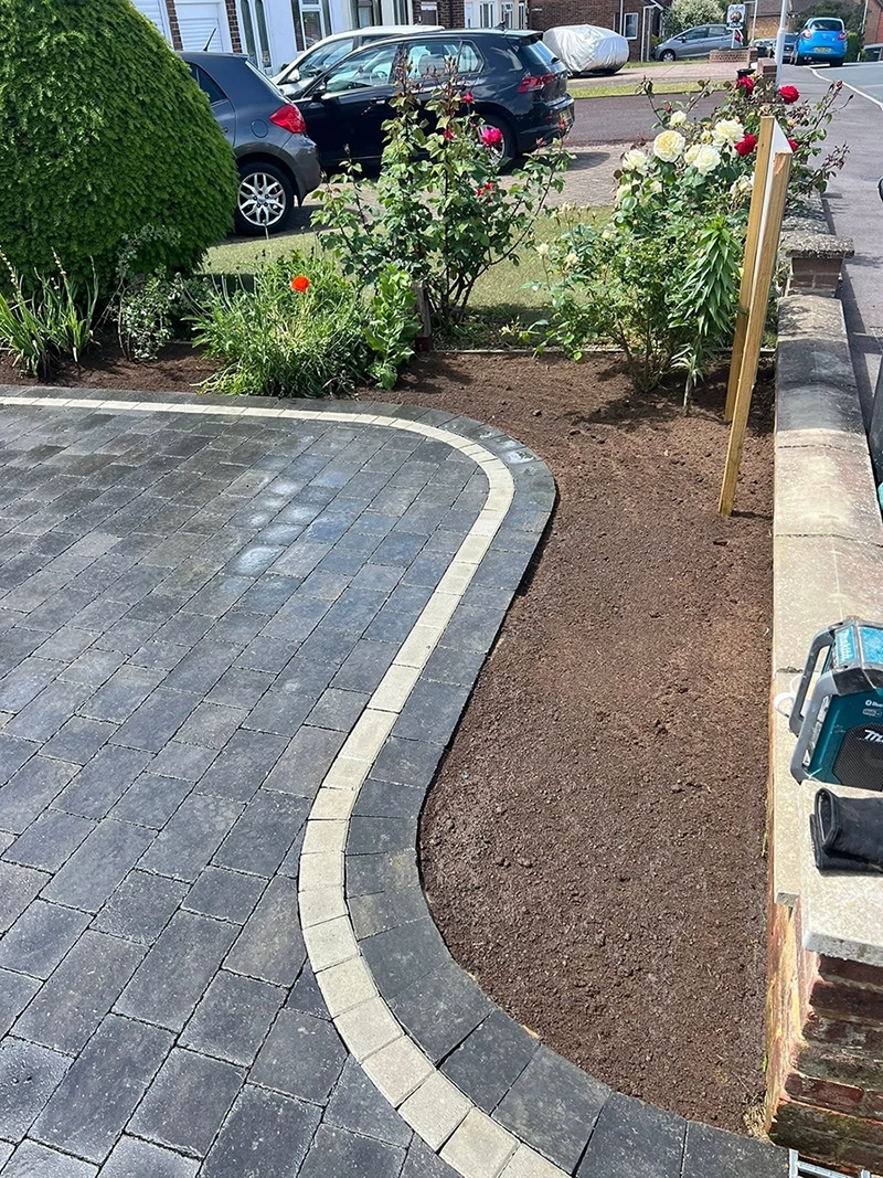 Curved edge patio with dark gray and beige paving stones next to freshly cleared soil bed bordered by low stone wall and a wooden post, with rose bushes and other plants in the background and cars parked nearby.