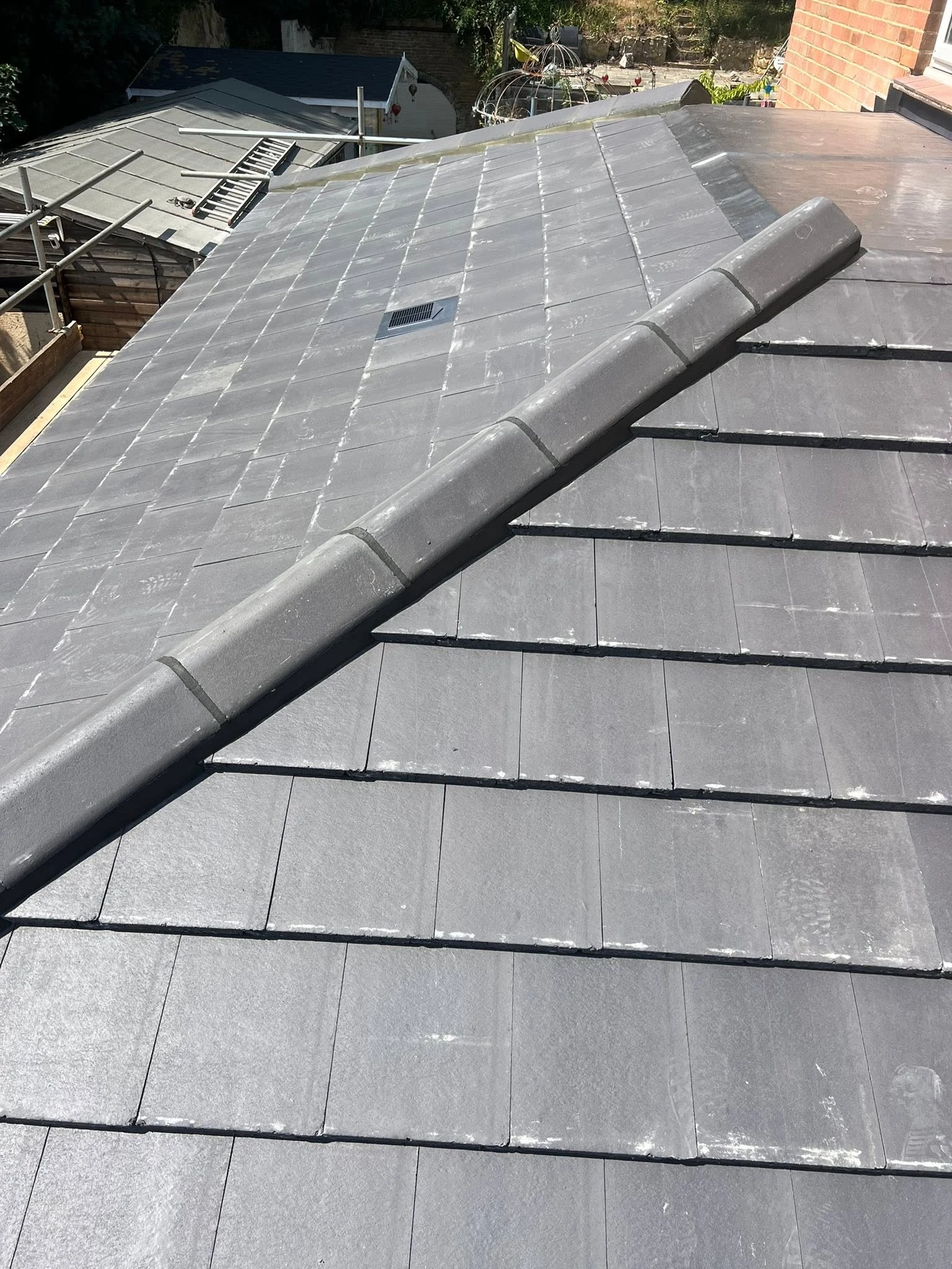 Close-up view of a gray tiled rooftop with two roof sections meeting at a ridge covered by ridge tiles, with a vent visible on the far section.