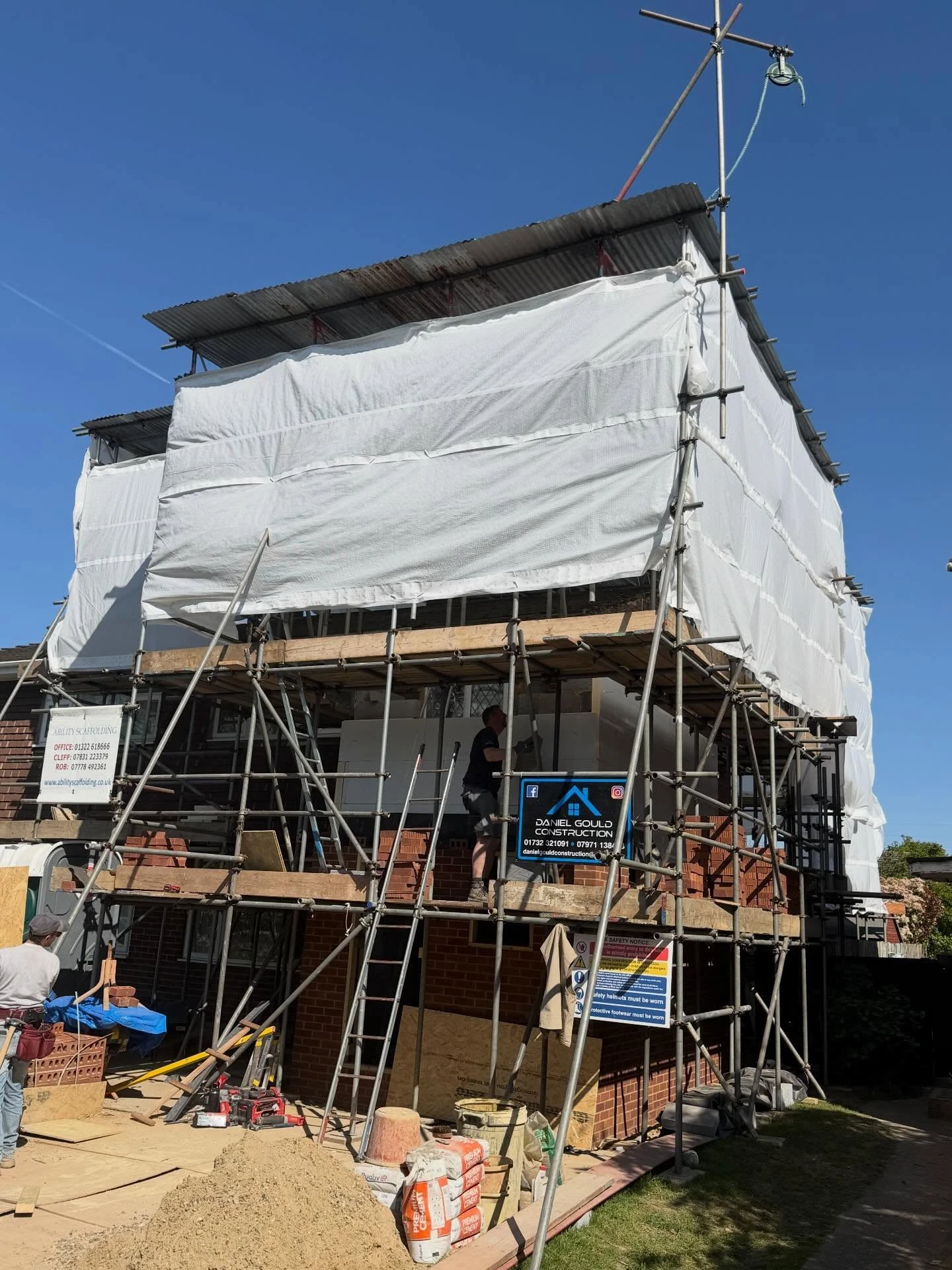 Brick house undergoing construction with scaffolding covered in white protective sheeting and two workers visible, one on the scaffolding and one on the ground, with construction materials and tools around the site under a clear blue sky.