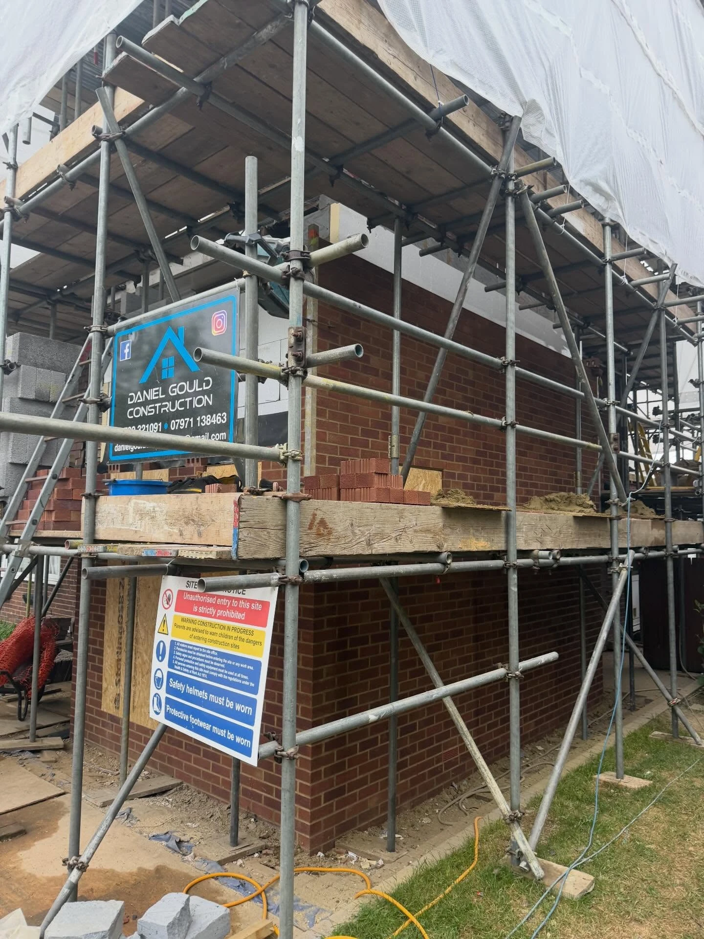 Construction site with scaffolding surrounding a brick building under renovation, featuring a sign for Daniel Gould Construction and safety warnings requiring helmets and protective footwear.