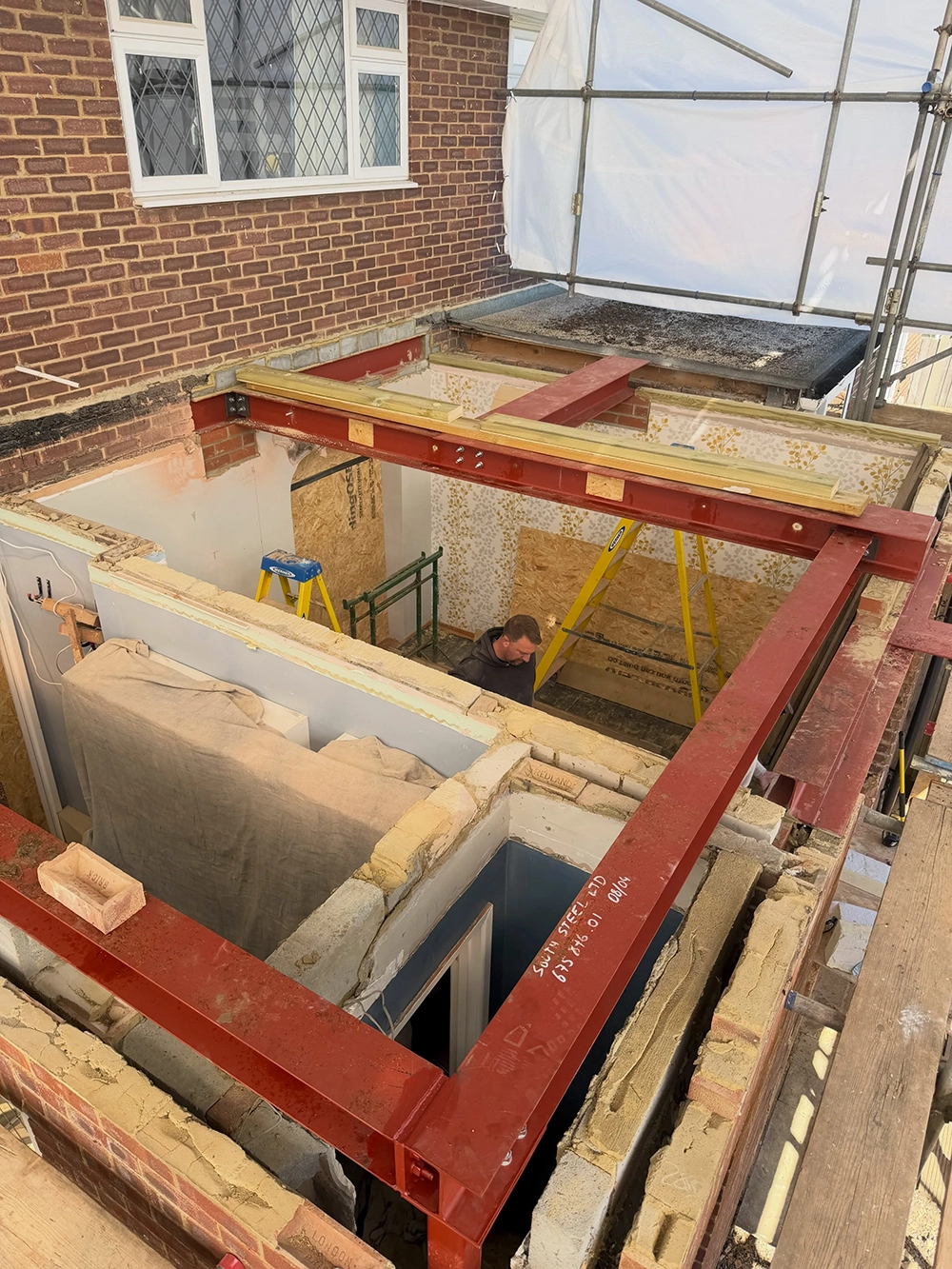 Construction site of a house extension with exposed brick walls, red metal support beams, a man working inside, ladders, scaffolding, and a covered section with a tarp.