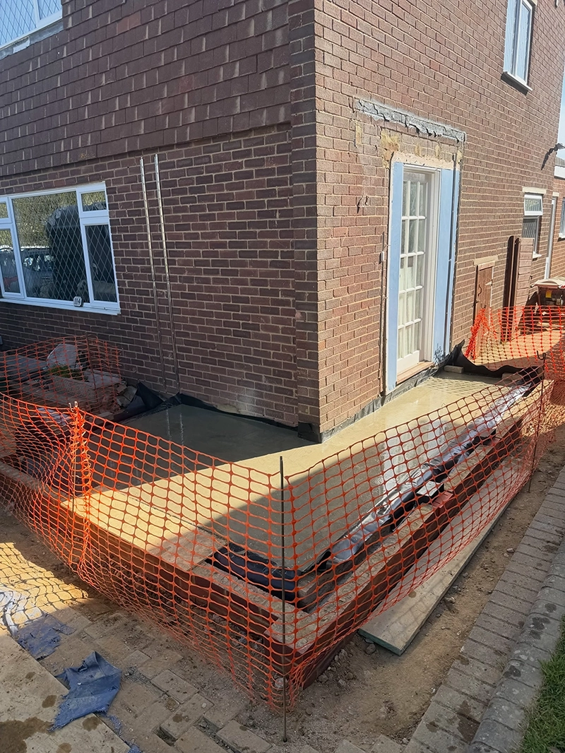 Newly poured concrete patio adjacent to a brick house with white-framed windows and a white door, surrounded by orange construction safety fencing and construction materials on the ground.