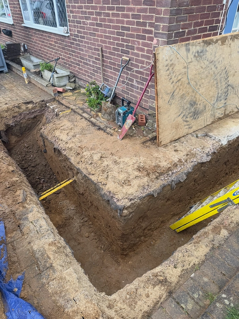 Deep rectangular trench dug in front of a brick house wall, with construction tools like shovels and a level leaning against the wall and a plywood sheet nearby.