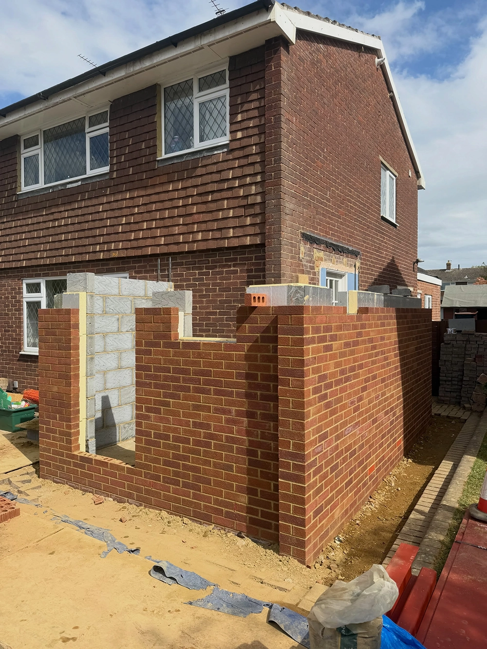 A brick house under renovation with a new brick extension under construction featuring unfinished walls and exposed concrete blocks on a sunny day.
